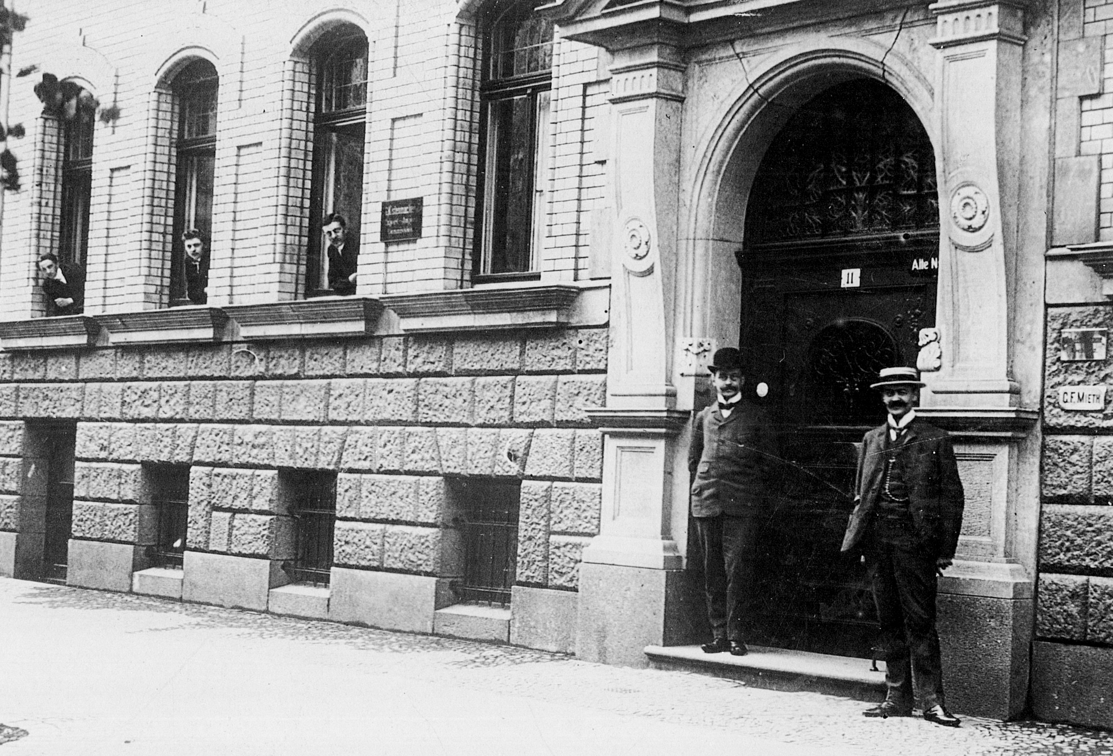 Blick von außen auf das erste Kistenmacher Büro. Ein gründerzeitliches Portal. Zwei Herren stehen vor der Tür. Weitere schauen im Erdgeschoss aus den Fenstern. Leipzig 1902